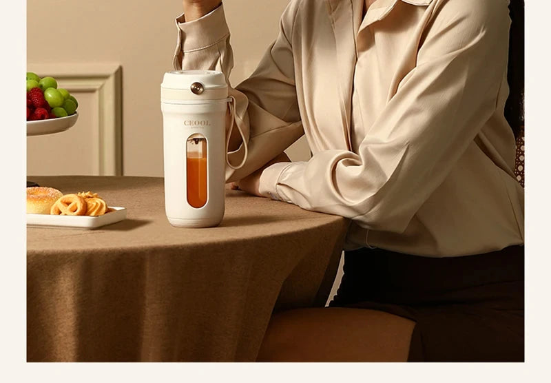 A portable white electric blender with a glass viewing window, sitting on a table next to a woman in a beige shirt.