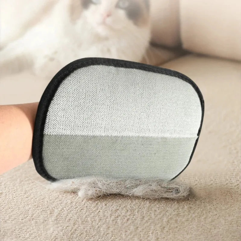 A hand wearing a two-tone grey pet hair removal mitt, lifting fur from a fabric sofa with a blurred cat nearby.