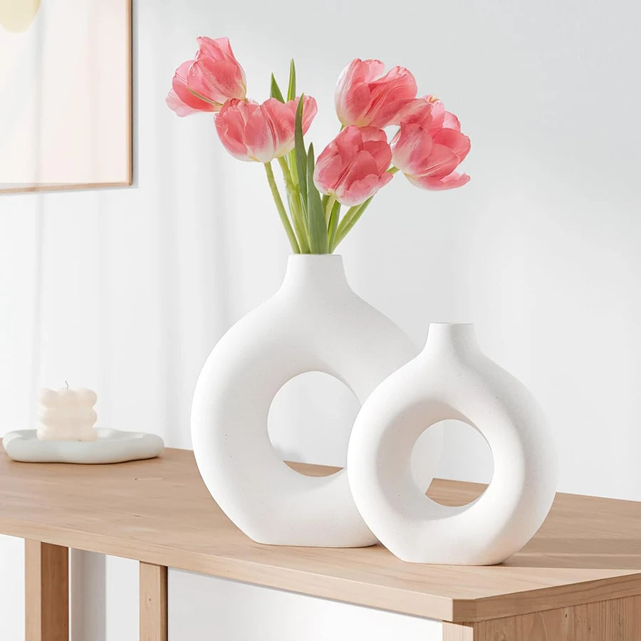 Two minimalist, white Nordic-style circular vases on a wooden table, with one holding a bouquet of pink tulips.