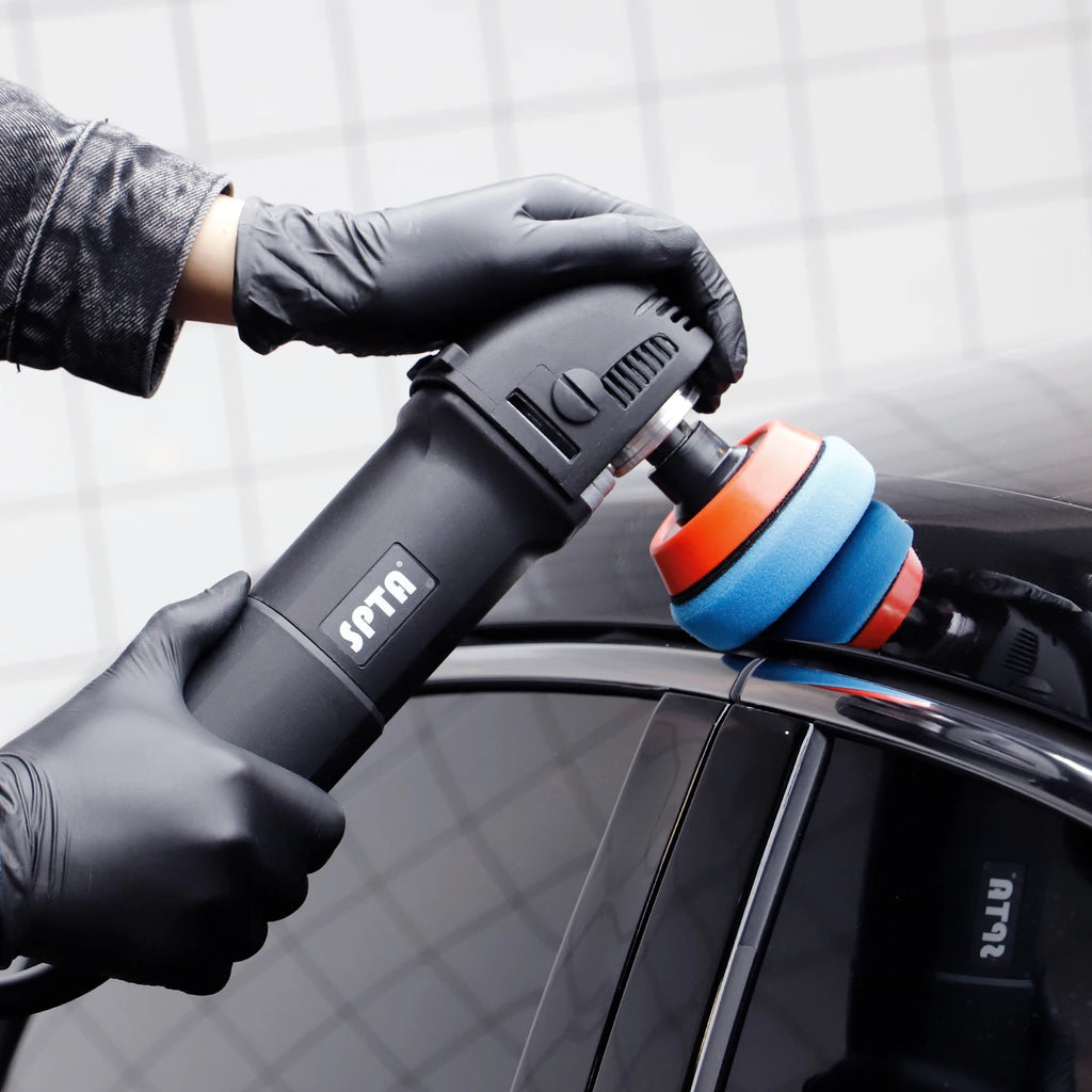 Gloved hands hold a black SPTA rotary drill fitted with round blue and orange foam polishing pads on a car.