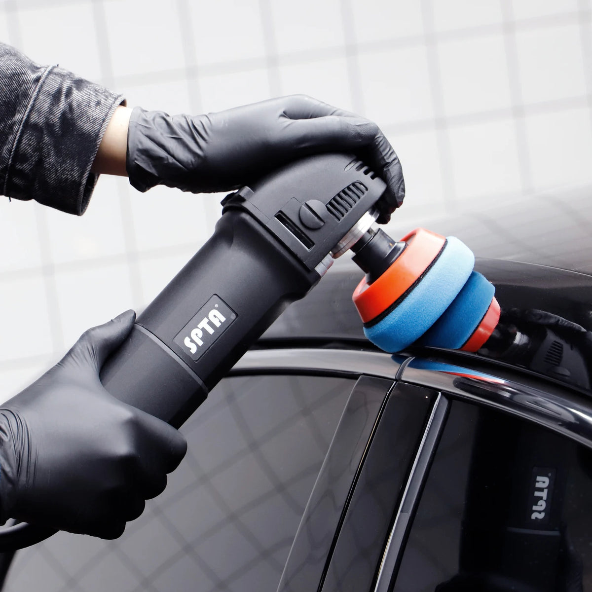 Gloved hands hold a black SPTA rotary drill fitted with round blue and orange foam polishing pads on a car.