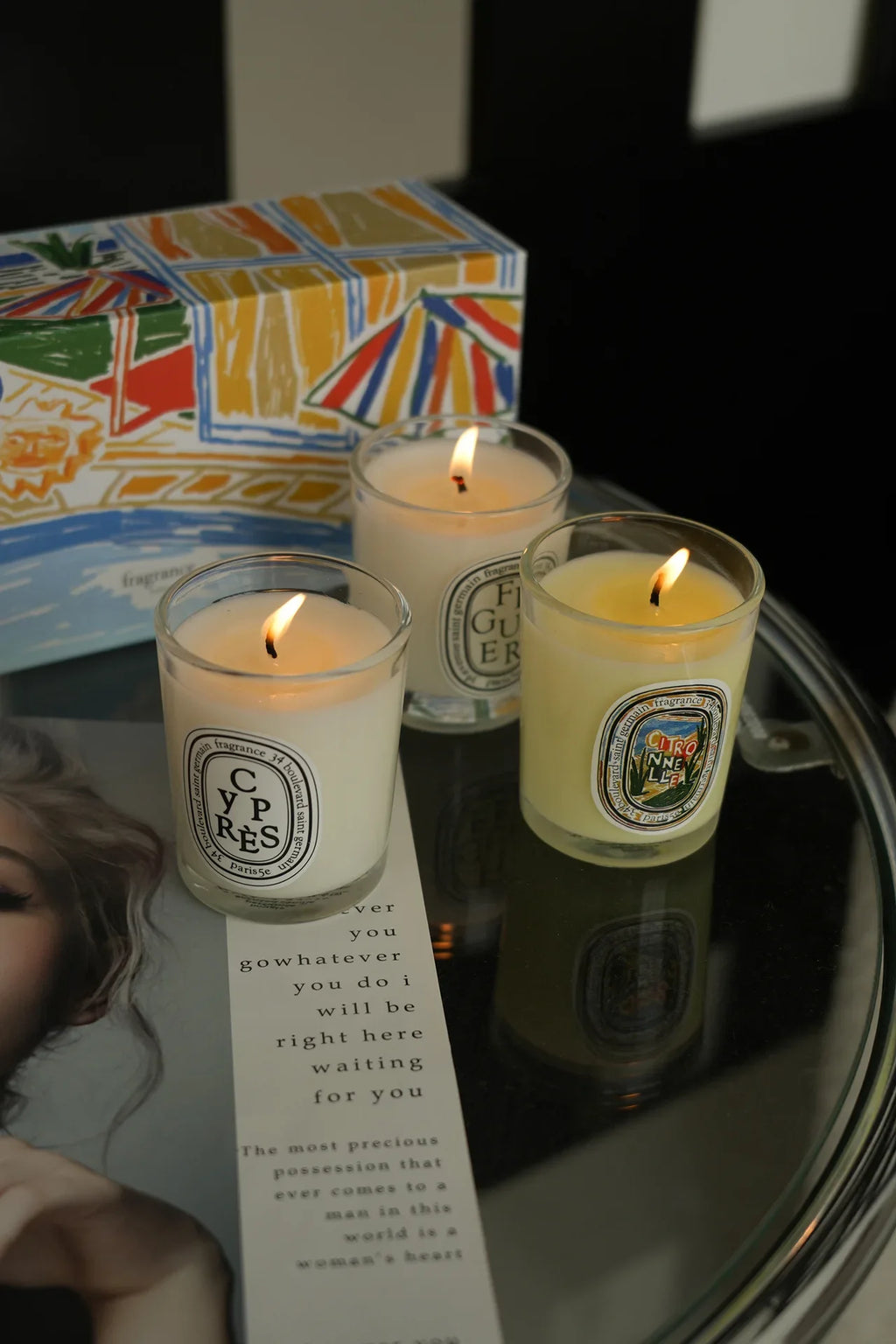 Three lit scented candles in glass jars with label designs, arranged on a table beside an illustrated gift box.
