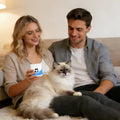 A couple sits on a rug with their fluffy cat, with the woman holding a white jar of cat taurine supplement.