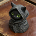 A small black cat figurine wearing an intricately carved hooded robe, featuring striking, bright green eyes.