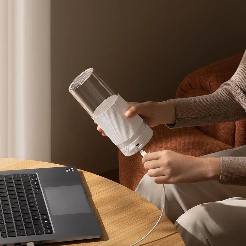 A person charging a white, minimalist portable blender with a clear glass top using a USB cable next to a laptop.