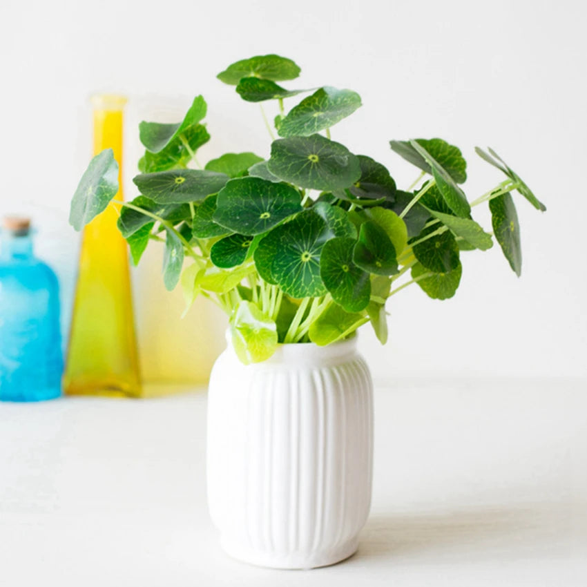 Artificial lotus leaves in a ribbed white ceramic pot, with blurred yellow and blue glass bottles in the background.