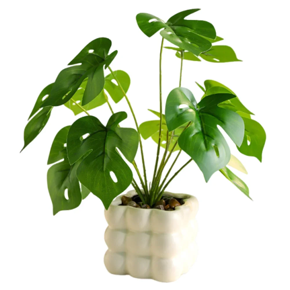 Artificial monstera plant with green split leaves in a decorative, white, textured ceramic pot with pebble soil.