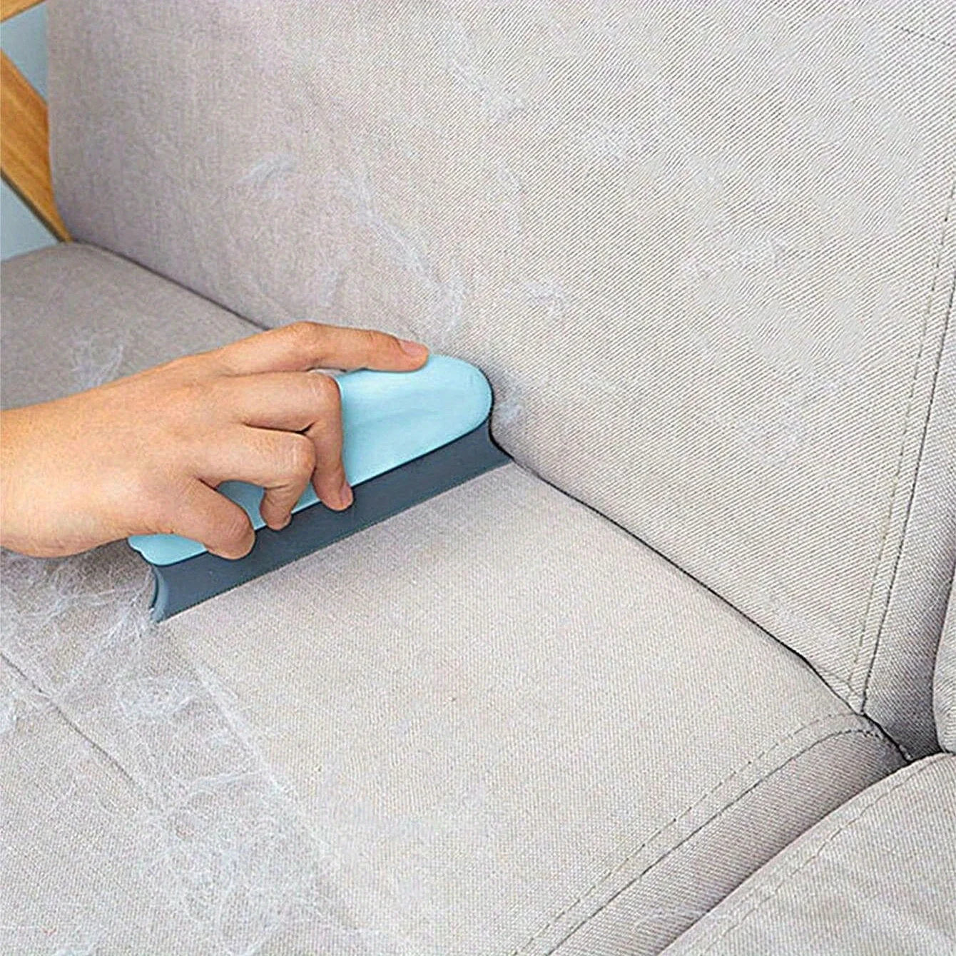 A hand using a light blue and grey lint remover tool to clean pet hair from a beige fabric sofa.