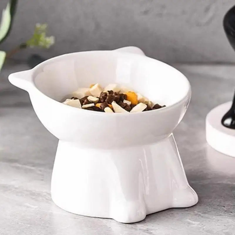 White ceramic cat-shaped elevated pet bowl with a 15-degree tilt, filled with dry food, on a grey surface.