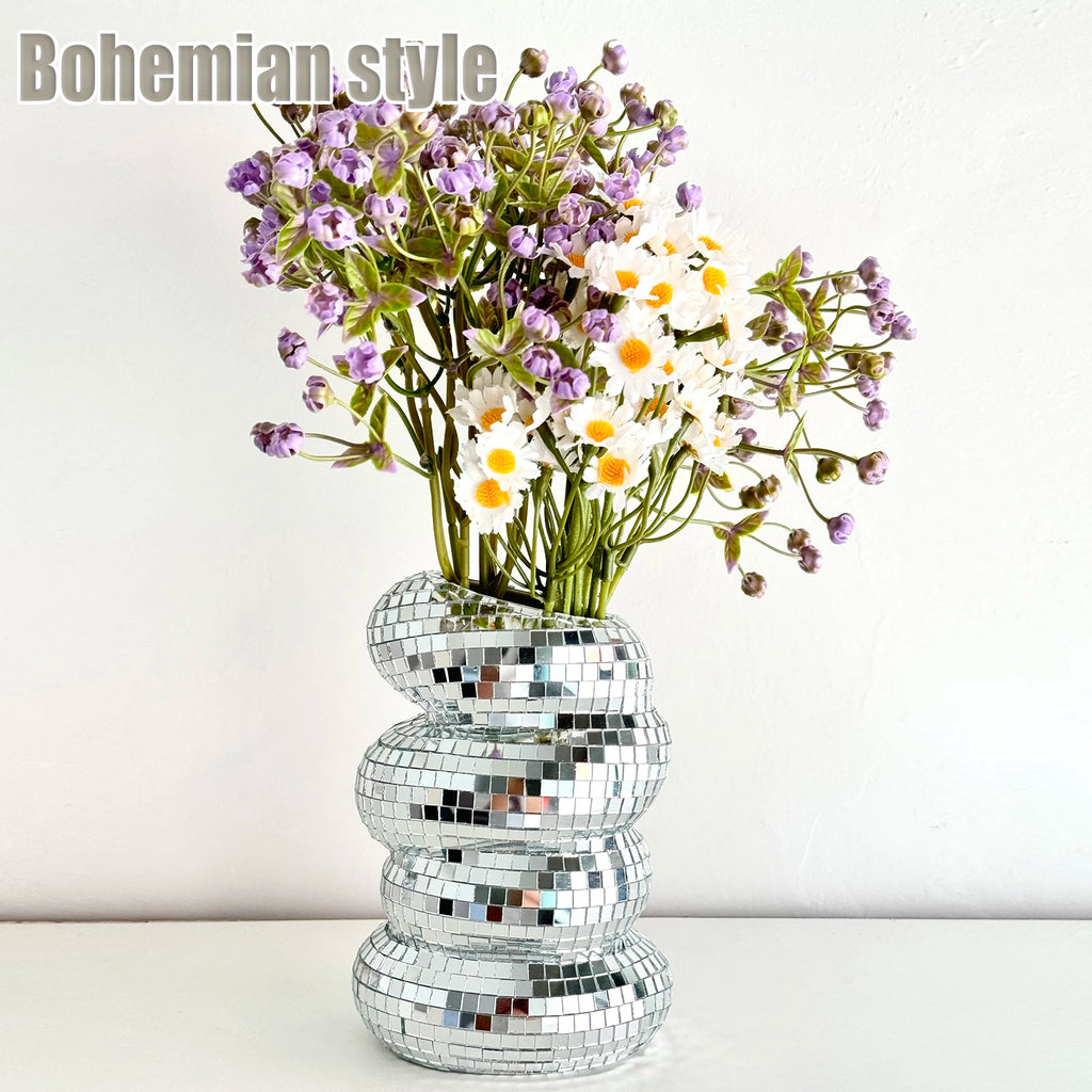 A wavy-shaped silver mirror disco ball vase, filled with purple flowers and white daisies on a white surface.
