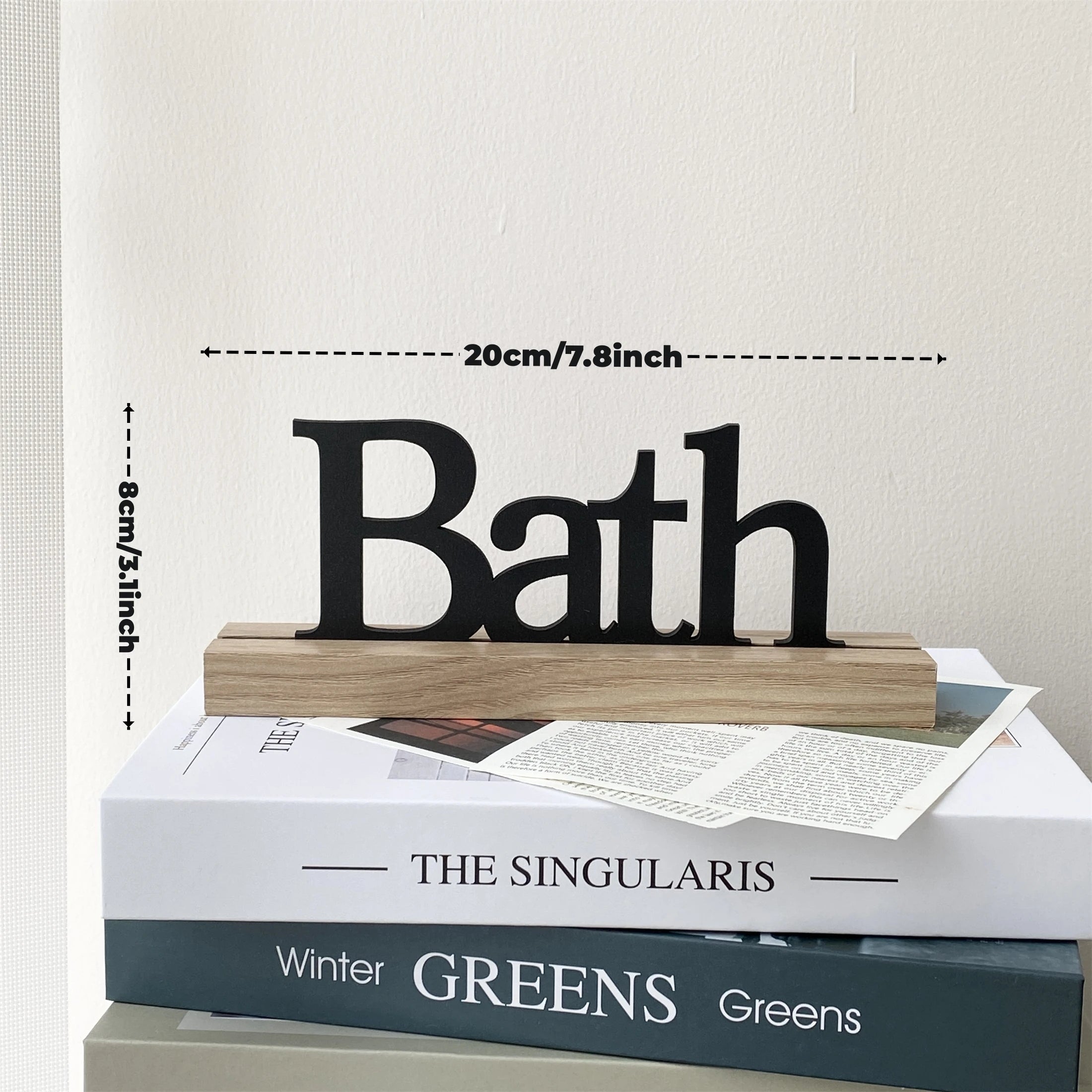 A rustic black metal 'Bath' sign standing on a natural light wood base, resting on top of stacked decorative books.