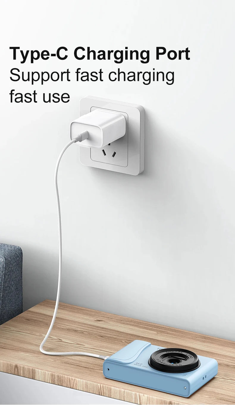 A light blue plastic digital camera charging via a white USB Type-C cable plugged into a white wall power adapter.