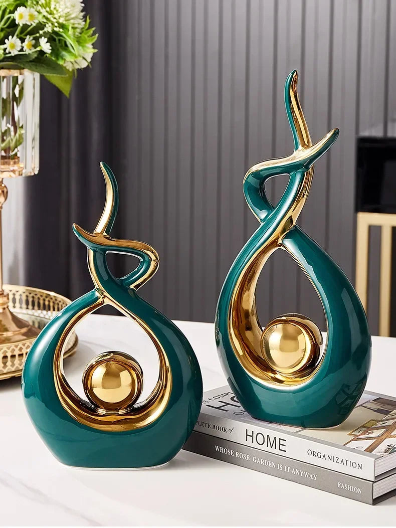 Two modern, abstract teal and gold ceramic sculptures with a smooth, glossy finish, standing on a surface.
