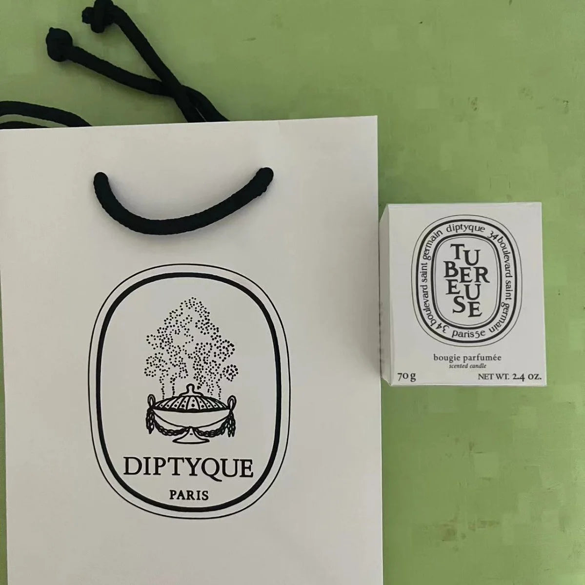 White 70g scented candle in box next to a matching white paper gift bag with black handles and branding.