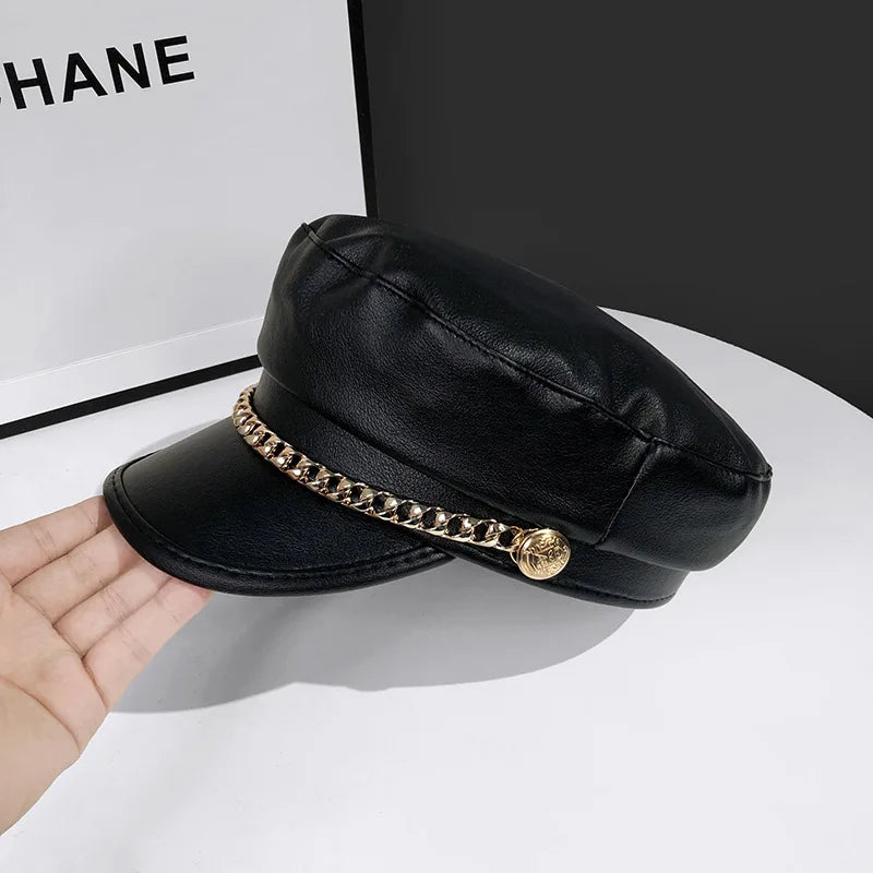 A stylish black faux-leather baker boy cap featuring a decorative gold chain and side button detail.