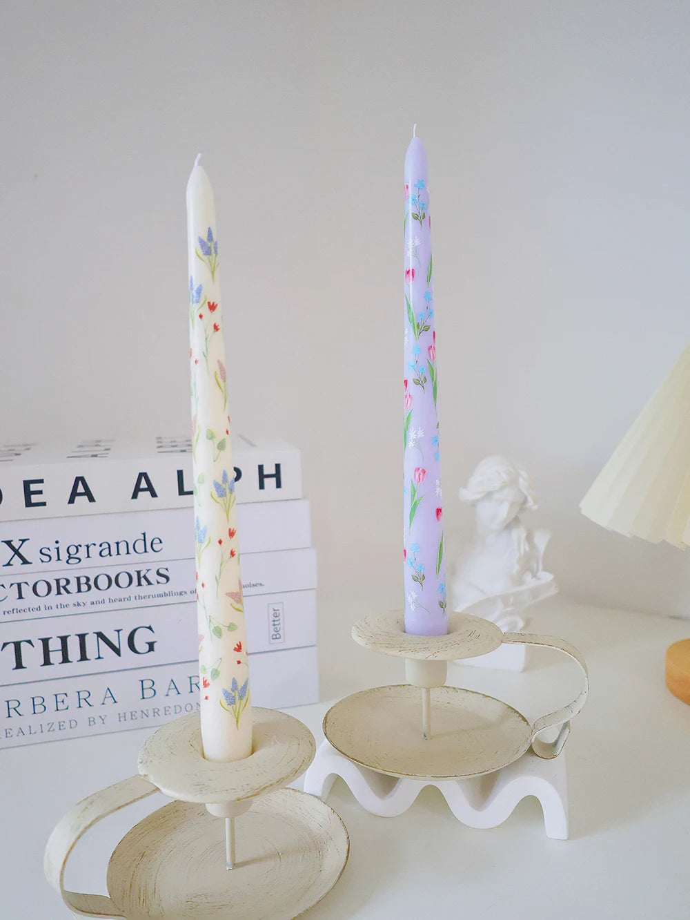 Two taper candles with floral patterns, one cream and one lavender, set in vintage-style metal candle holders.