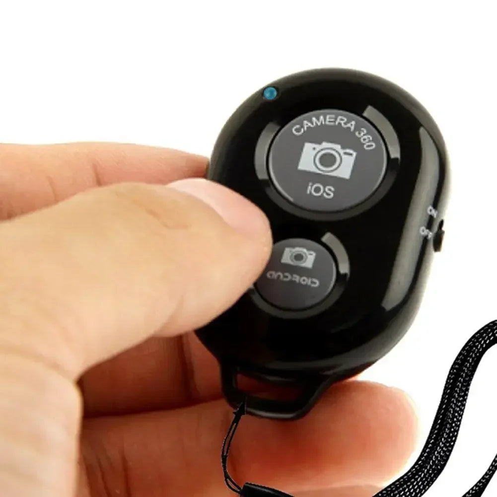 Black plastic Bluetooth camera remote shutter with iOS and Android buttons and a wrist strap held in a hand.
