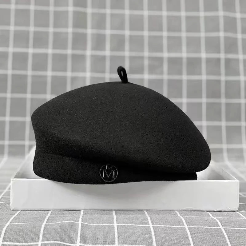 Classic black wool felt beret with a small top tab and circular metal charm detail on the side.