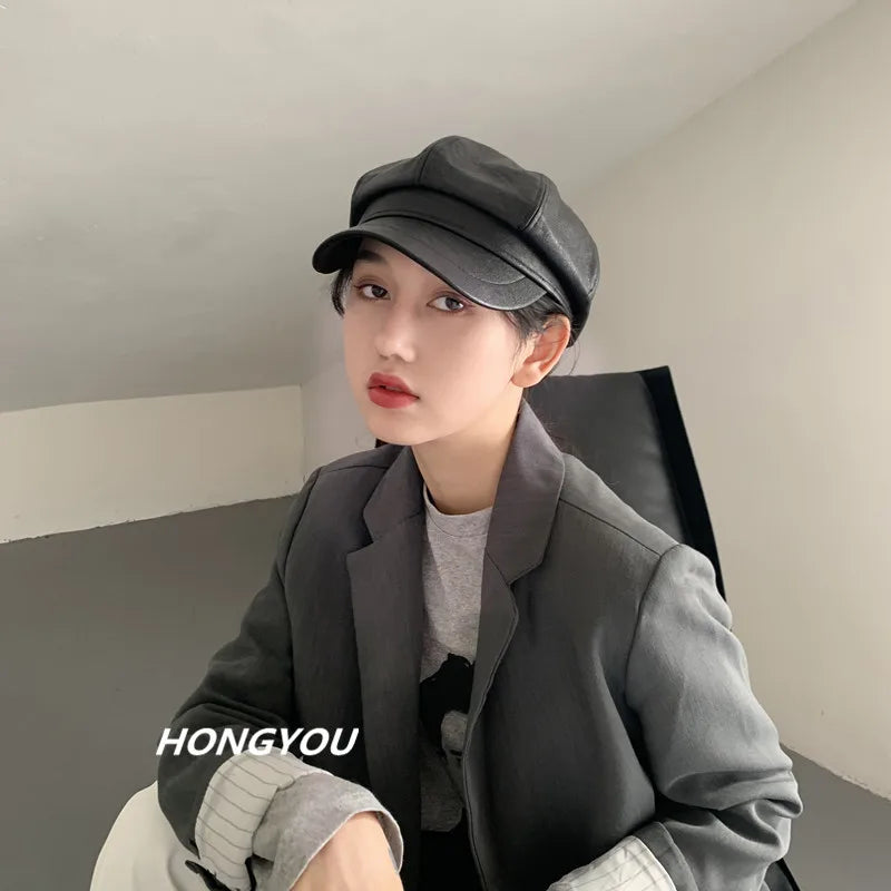 A stylish black faux-leather baker boy cap with a short peak, worn by a woman in a grey blazer.
