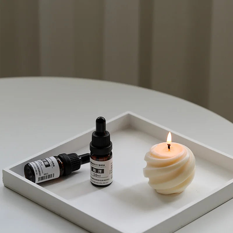 A white tray holding two small amber dropper bottles and a lit, cream-coloured swirled spherical candle.