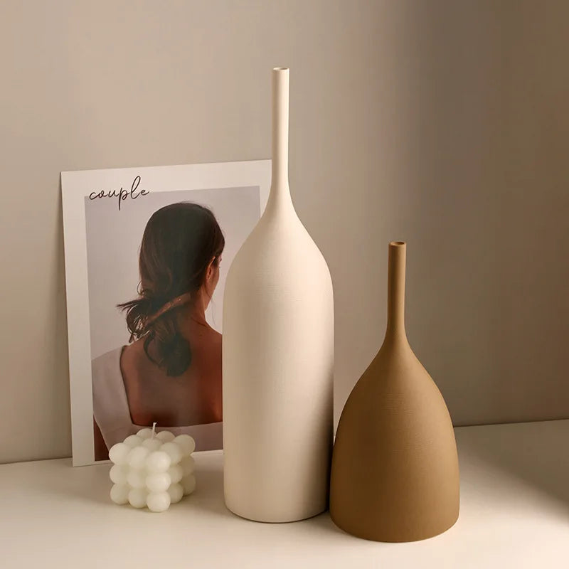Two minimalist, handmade ceramic vases in cream and tan with slim necks, set beside a bubble candle and art print.