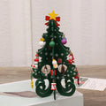 A dark green wooden Christmas tree ornament decorated with hanging colourful baubles and festive figures, topped with a star.