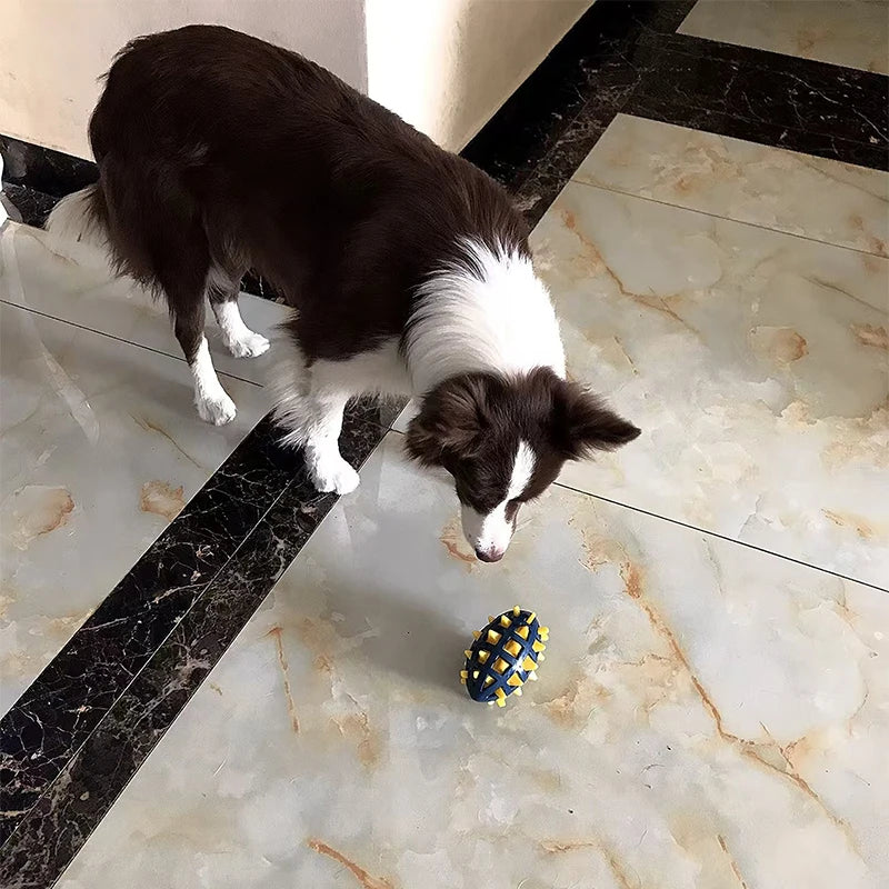 A dog looking at a durable, blue and yellow rubber rugby ball toy with a spiked, textured design on a tiled floor.