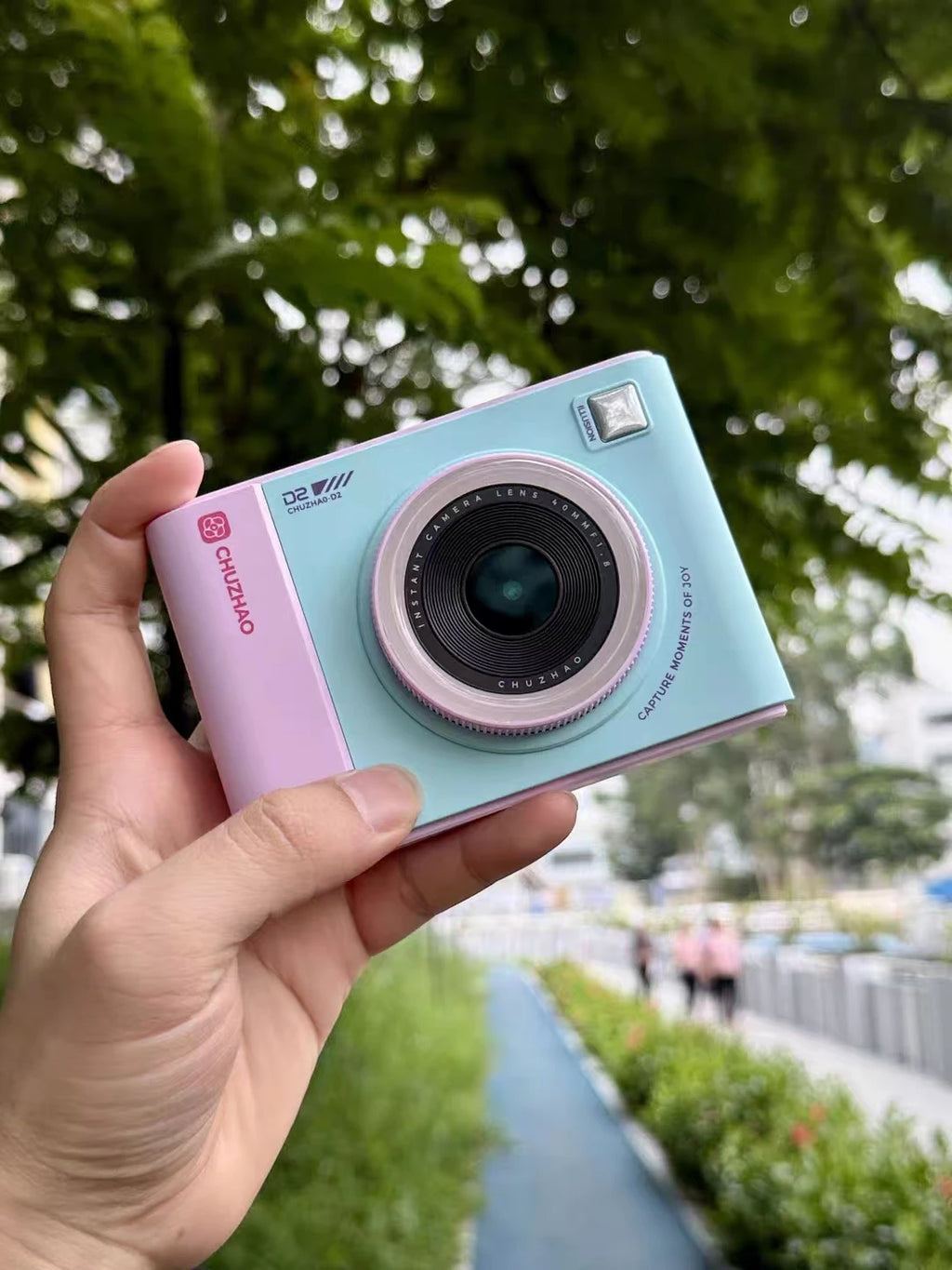 Pastel blue and pink CHUZHAO D2 instant camera held in a hand against a blurred outdoor background.