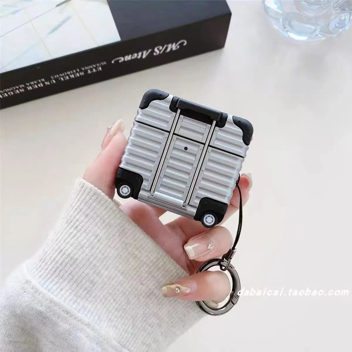 Silver suitcase-style AirPods case with a keychain, featuring a rigid, textured design and black accents.