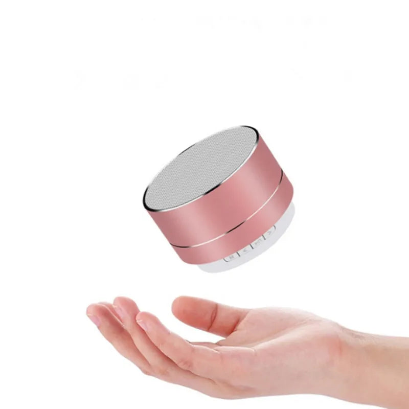 Compact, rose gold aluminium portable Bluetooth speaker hovering above a hand. Includes a built-in LED and subwoofer.