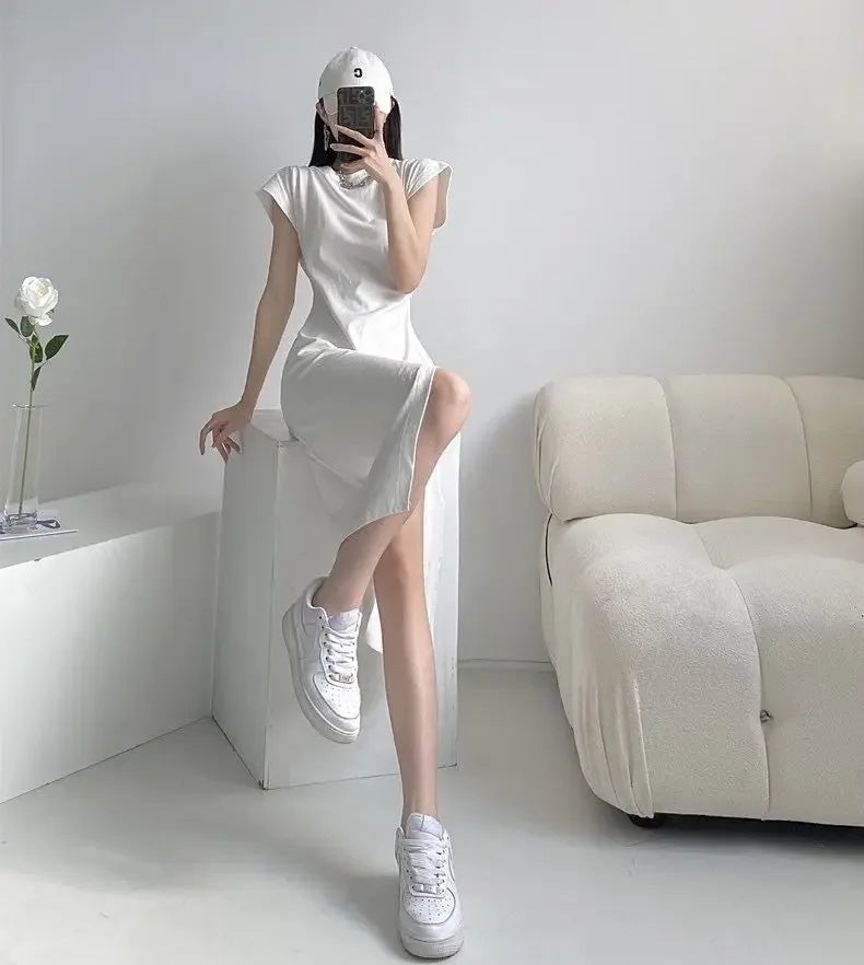 A woman wearing a white cap-sleeve midi dress with a side slit, styled with white trainers and a baseball cap.