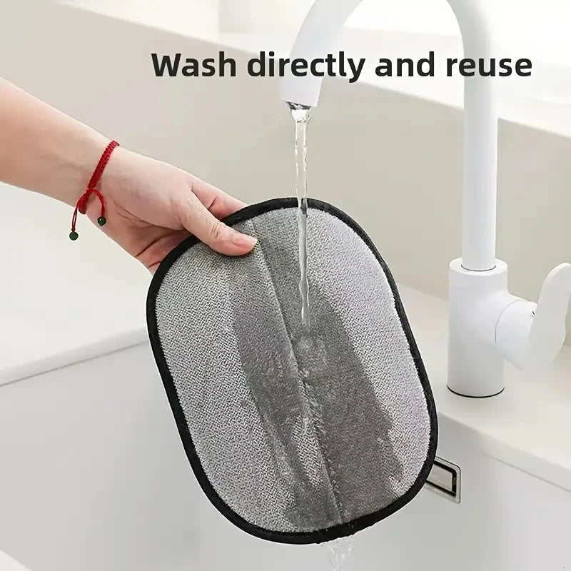A hand holds a grey, oval-shaped reusable cleaning pad with a black trim under a running kitchen tap.