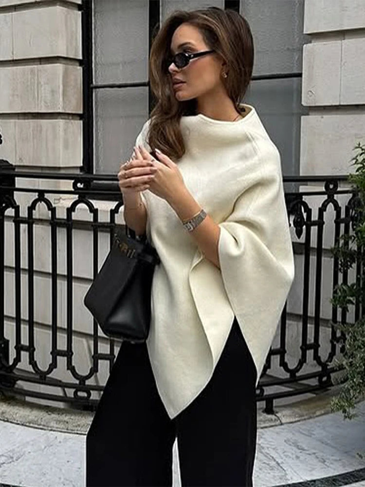 Woman wearing an off-white knitted cape jumper with an asymmetrical draped hem and high neckline.