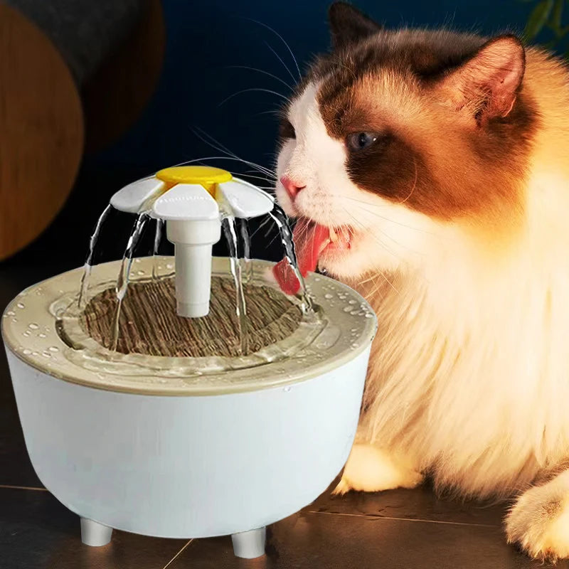 A fluffy cat drinks from a white, round pet water fountain featuring a yellow and white daisy-shaped flower spout.