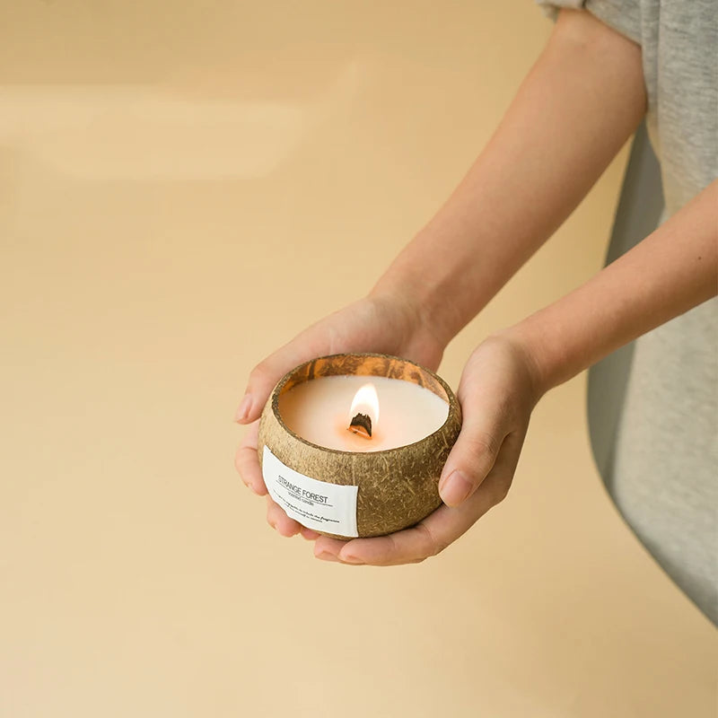 Lit scented candle in a textured coconut shell bowl held in hands against a neutral background.