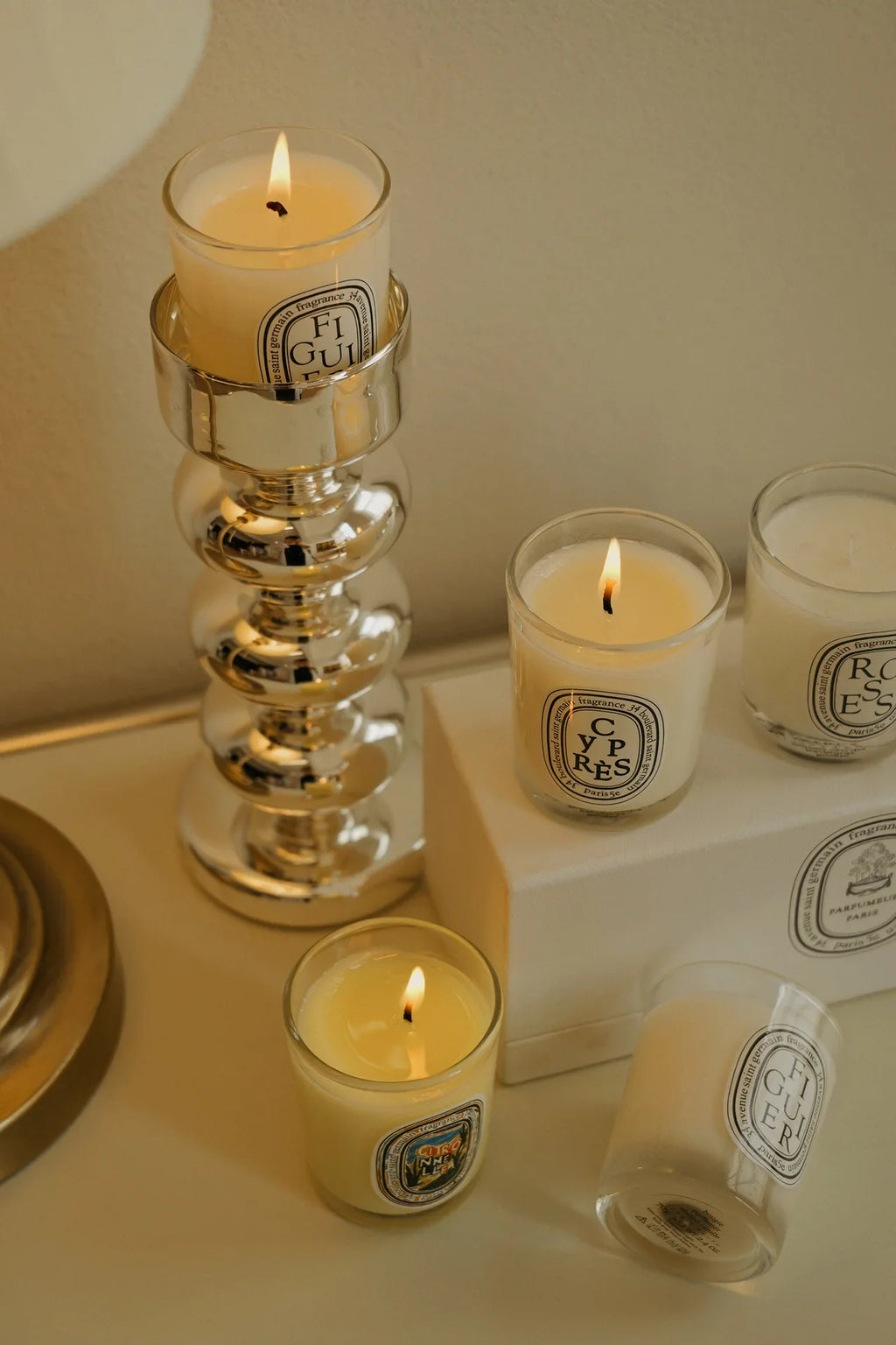 Three lit scented candles in glass jars, one in a silver pedestal holder, and one laid on its side on a white box.