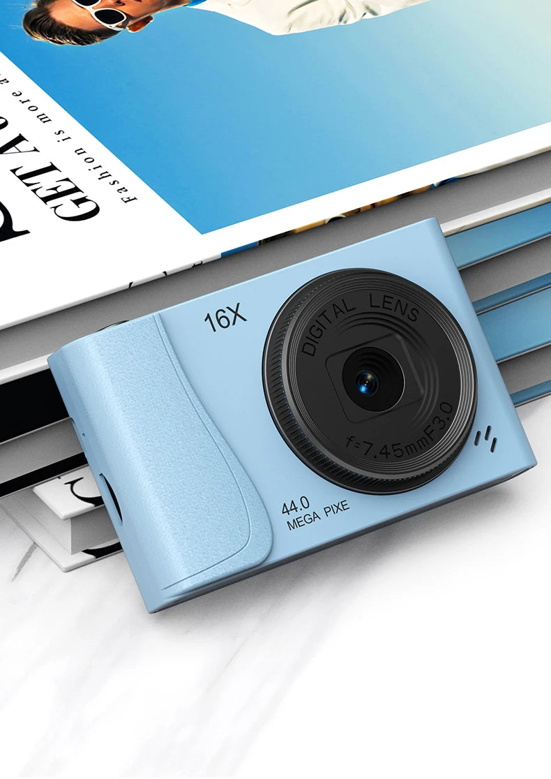 Light blue plastic digital camera with a 16x zoom lens and 44-megapixel sensor, resting on top of a stack of books.