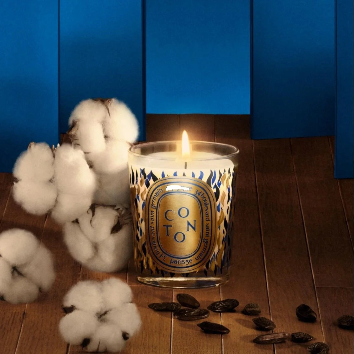 Lit candle in a decorative glass jar with blue and gold patterns, surrounded by white cotton and dark cocoa beans.