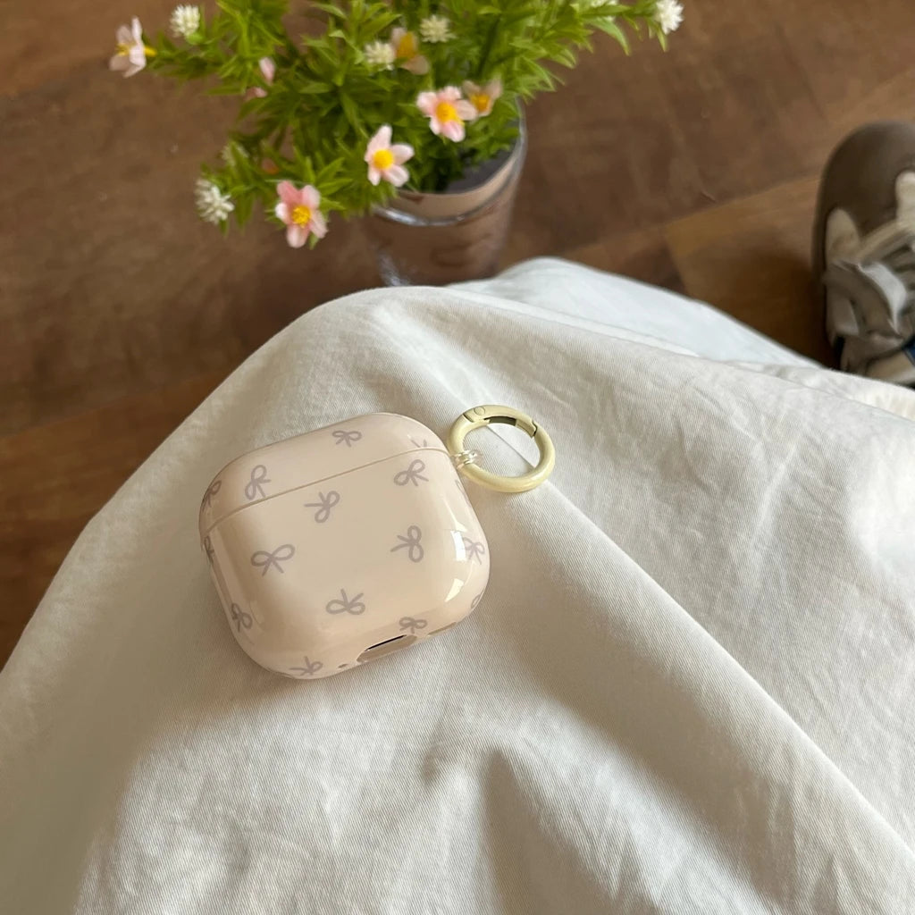 Glossy pale pink AirPods case with a grey bow pattern and a matching ring clip on white fabric.