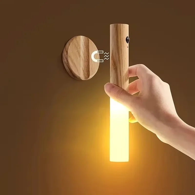 A hand detaches a sleek, wooden motion-sensor LED night light from its magnetic wall mount, emitting a warm glow.