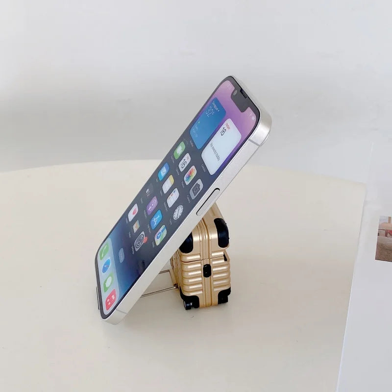 Gold suitcase-style protective case for AirPods, doubling as a smartphone stand on a white surface.