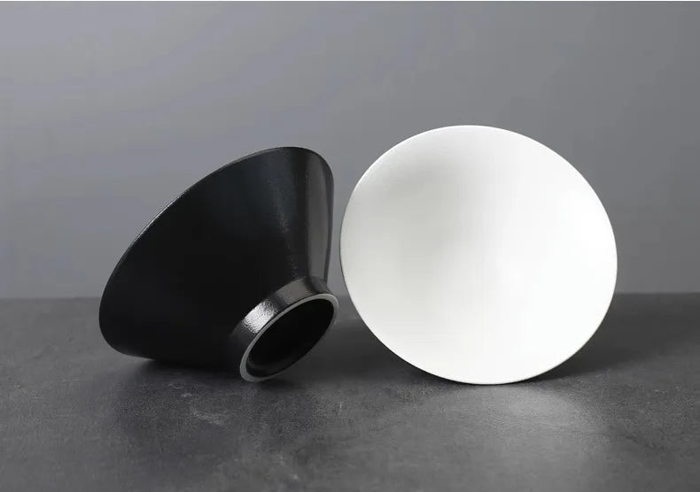 Two minimalist ceramic bowls in matte black and white, featuring a sleek, wide-brimmed conical design.