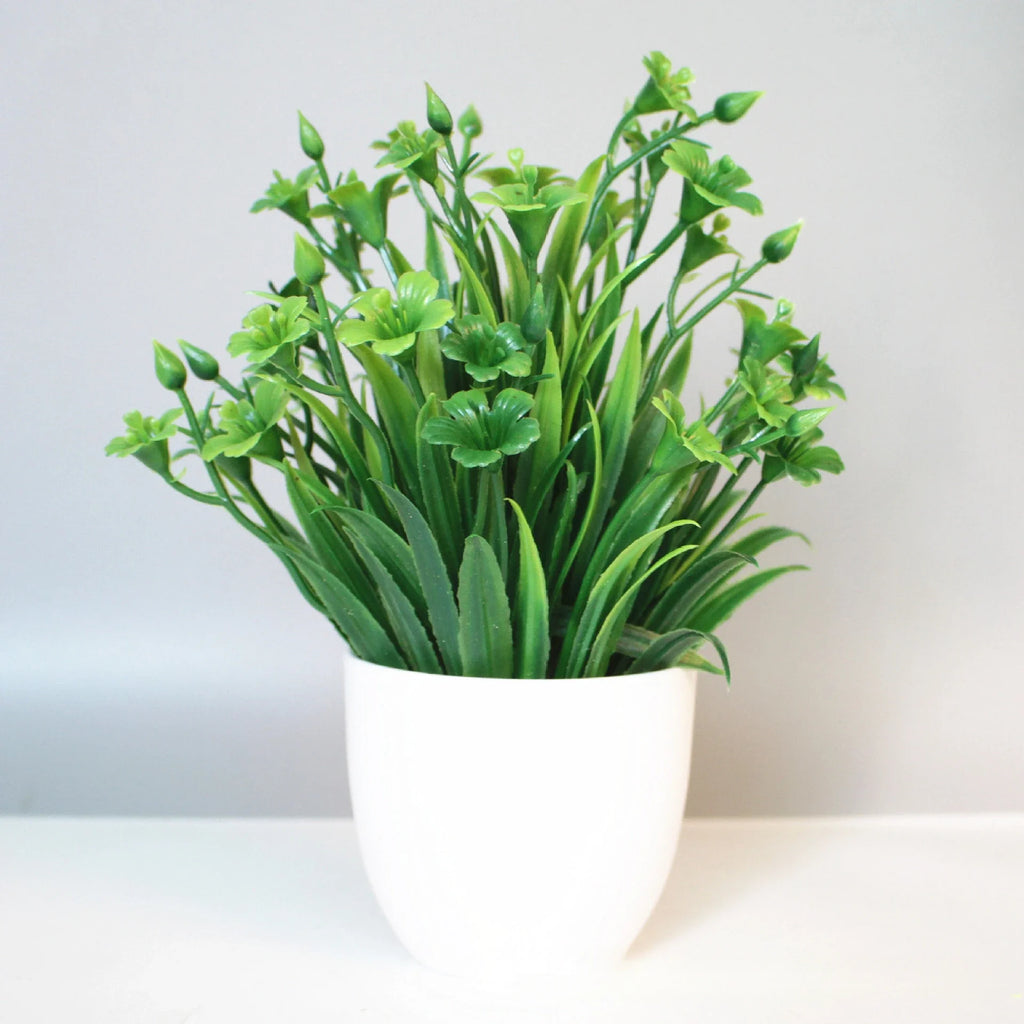 Artificial green plant with small flowers and buds in a simple white pot against a light grey background.