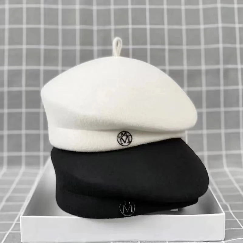 Two classic wool felt berets, one white and one black, each featuring a small, circular metallic 'M' emblem.
