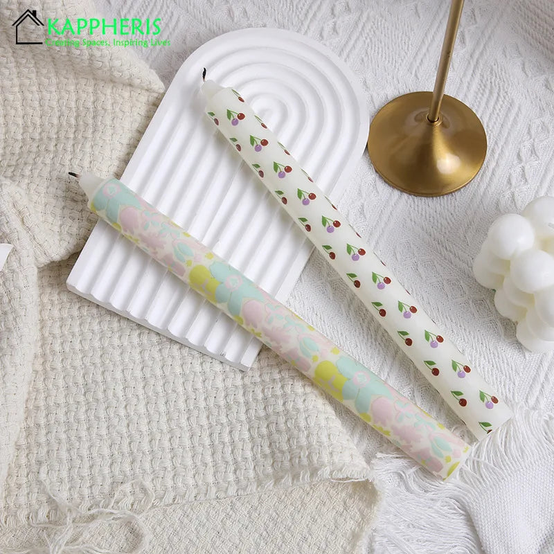 Two taper candles: one with a floral pastel print and one with a small cherry pattern, set on a white wavy tray.