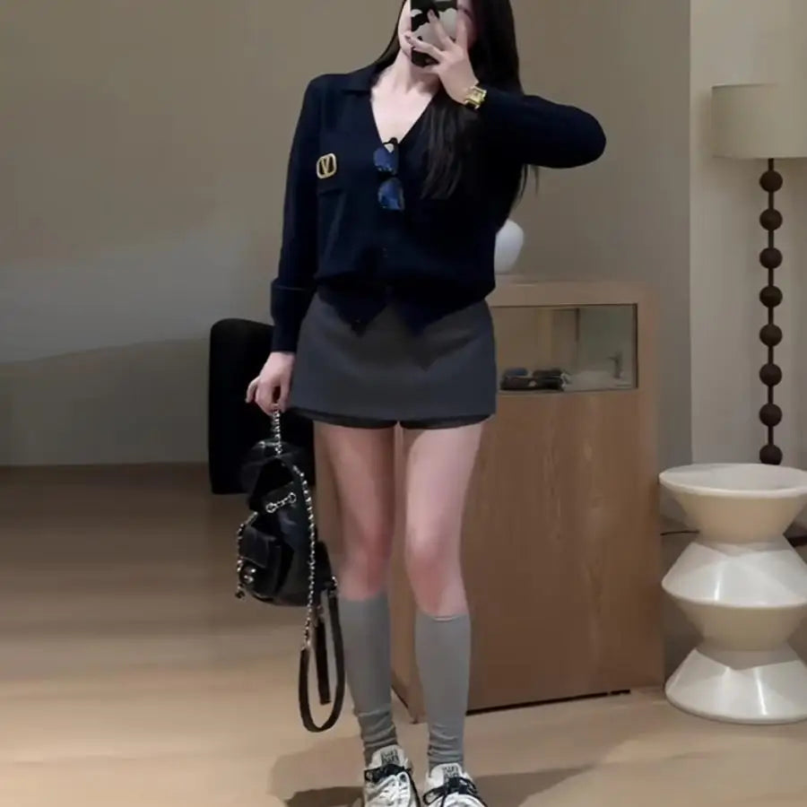 A woman wearing a black V-neck cardigan, grey mini skirt, grey knee-high socks, and carrying a black chain strap bag.