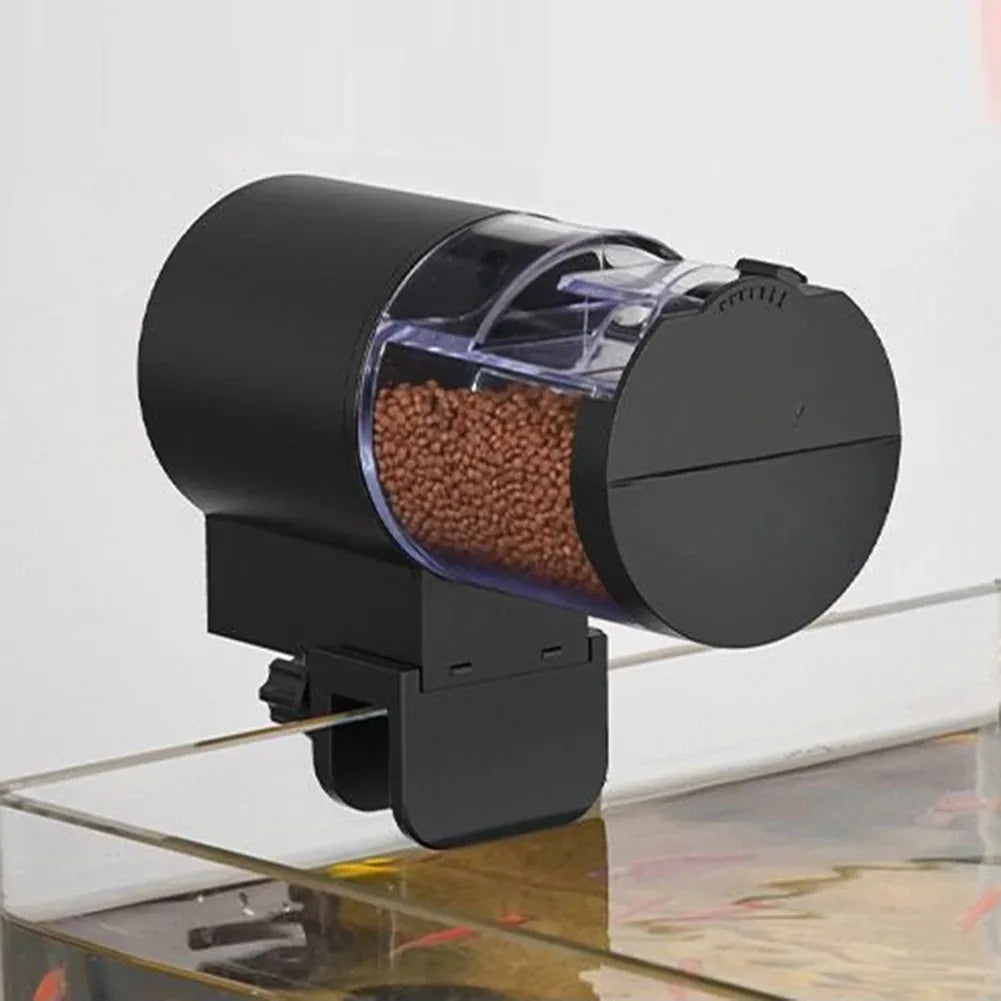 Black automatic fish feeder with a clear, cylindrical food container filled with brown pellets, mounted on a tank rim.