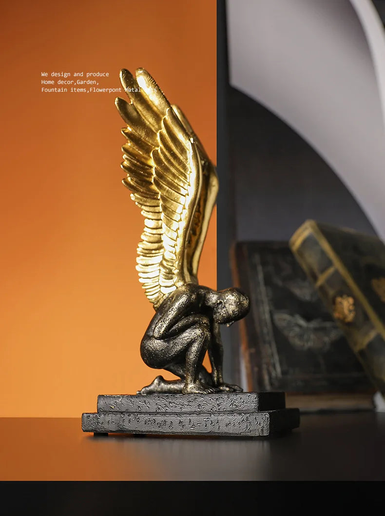 A kneeling angel sculpture with textured dark grey body and shimmering metallic gold wings on a rectangular base.