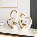 Two modern minimalist ceramic heart ornaments in white with polished gold detailing, displayed on a marble table.