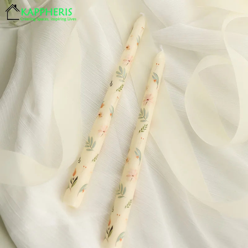 Two cream taper candles decorated with delicate floral and sage leaf patterns, resting on soft white fabric.